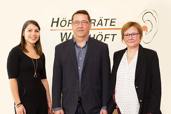 Drei Mitarbeitende eines Hörgeräteakustikers stehen vor dem Firmenlogo im Geschäft.